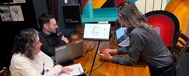 Picture of Heather, Brian and Allen gathered around a table with various laptops and monitors, collaborating in a friendly atmosphere.